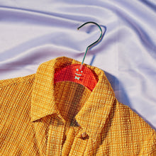 Load image into Gallery viewer, View of a yellow and orange plaid top on an orange hanger that has a green smiley face in the middle of it laying on light purple shiny fabric.
