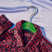 Load image into Gallery viewer, View of a red and black cheetah print top on a green hanger with a purple smiley face in the middle laying on a light purple shiny fabric.
