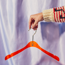 Load image into Gallery viewer, A hand coming out of a Cowichan sweater with a music note on the sleeve holding the hook of an orange acrylic hanger with a blue smiley face and bowtie on it in front of a light purple shiny curtain.
