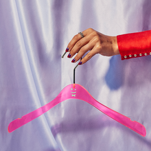Load image into Gallery viewer, A hand in a red sleeve with lots of buttons on the cuff holding a pink acrylic hanger with a green smiley face and bowtie printed on it, in front of a light purple shiny curtain.
