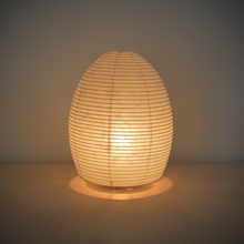 Load image into Gallery viewer, Image of an illuminated egg shaped paper lamp giving off a warm yellowish light.
