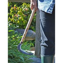 Load image into Gallery viewer, Image of a person in gardening clothes holding an extra-long wooden handled, hand-held sickle with a rough-hewn blade by their side with plants in the background.
