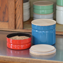 Load image into Gallery viewer, A small red container open with its open top sitting to the right in front of a larger blue container with a wooden top closed on a shelf, behind them an open cabinet with green and white containers stacked.
