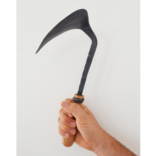 Load image into Gallery viewer, Side view of a hand holding a rough hewn, hand-hammered metal hook shaped blade with a wooden handle.
