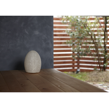 Load image into Gallery viewer, Image of a non-illuminated egg shaped paper lamp on the edge of a porch by a concrete wall with a fence and tree in the near distance.
