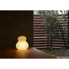 Load image into Gallery viewer, View of an illuminated mushroom shaped paper lantern with a yellowish glow on a porch with a fence and tree in the background.
