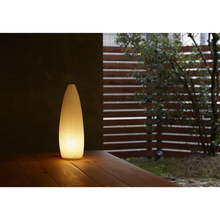 Load image into Gallery viewer, An illuminated simple LED torpedo-shaped paper lantern on the edge of a porch with a fence and tree in the background.
