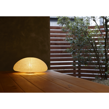 Load image into Gallery viewer, An illuminated simple LED low dome-shaped paper lantern on a porch with a fence and tree in the background.
