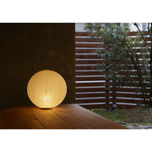 Load image into Gallery viewer, View of an illuminated simple ball-shaped LED paper lantern on a porch with a fence and tree in the background.

