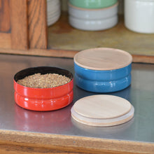 Load image into Gallery viewer, A small red container open with its open top sitting to the right in front of a small blue container with a wooden top closed on a shelf, behind them an open cabinet with green and white containers stacked.
